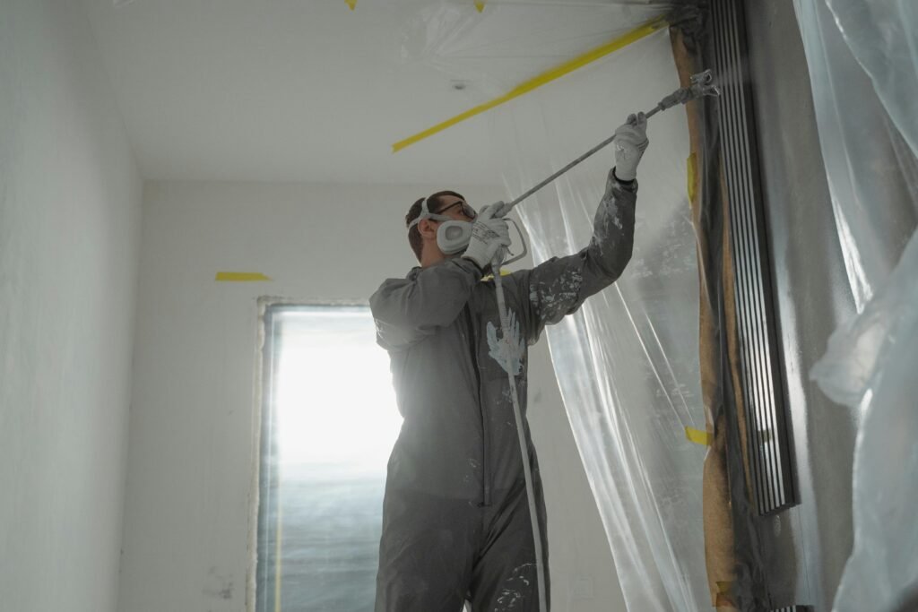 A professional painter wearing protective gear spray paints a wall indoors, working on a home renovation project.